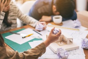 Group of architects looking stressed while working on an architectural model and shop drawing papers spread out on a table in an office.