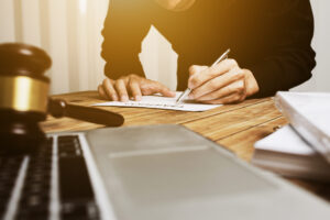 A man in a dark shirt signs a legal document with a pen at a wooden table next to a gavel, laptop, and folders.