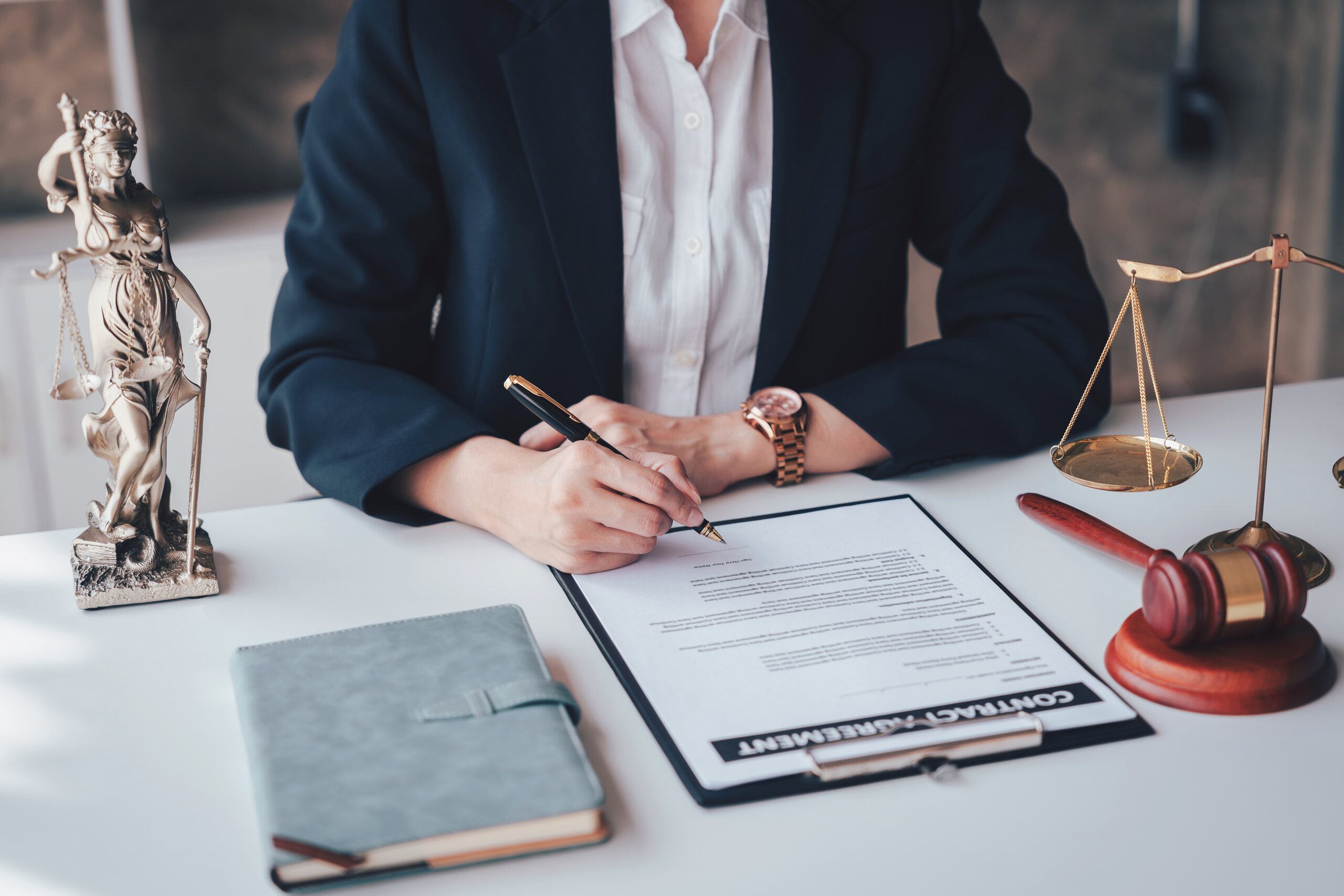 A lawyer in a dark suit sits at a white desk signing a document on a clipboard, surrounded by a Lady Justice statue, a wooden gavel, and legal scales, representing the probate court process for how to contest a will in Florida.