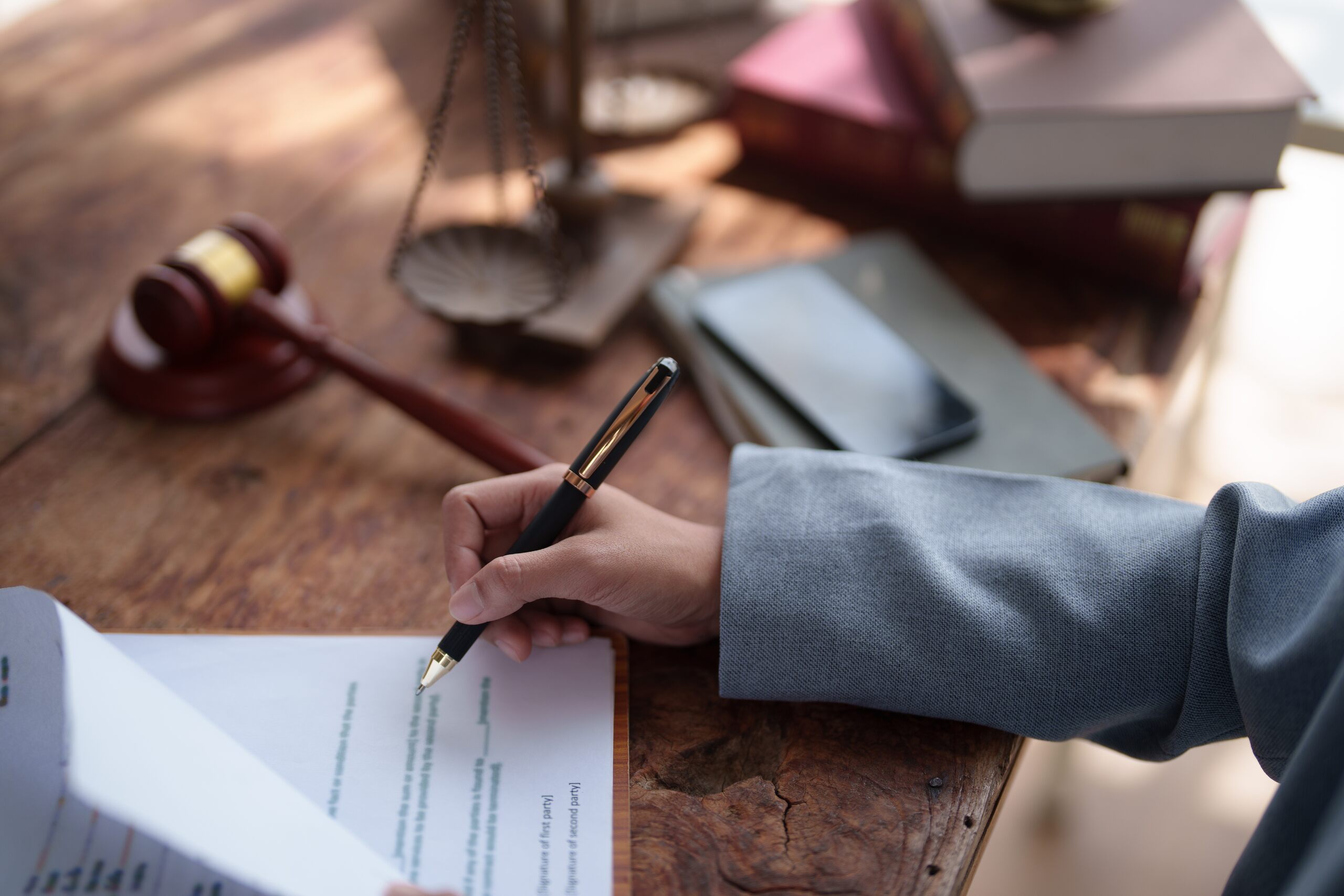 A close-up view of a person in a gray sleeve holding a pen and reviewing paperwork on a wooden table next to a gavel, legal scales, and law books, illustrating the collection of evidence for how to contest a will in Florida.
