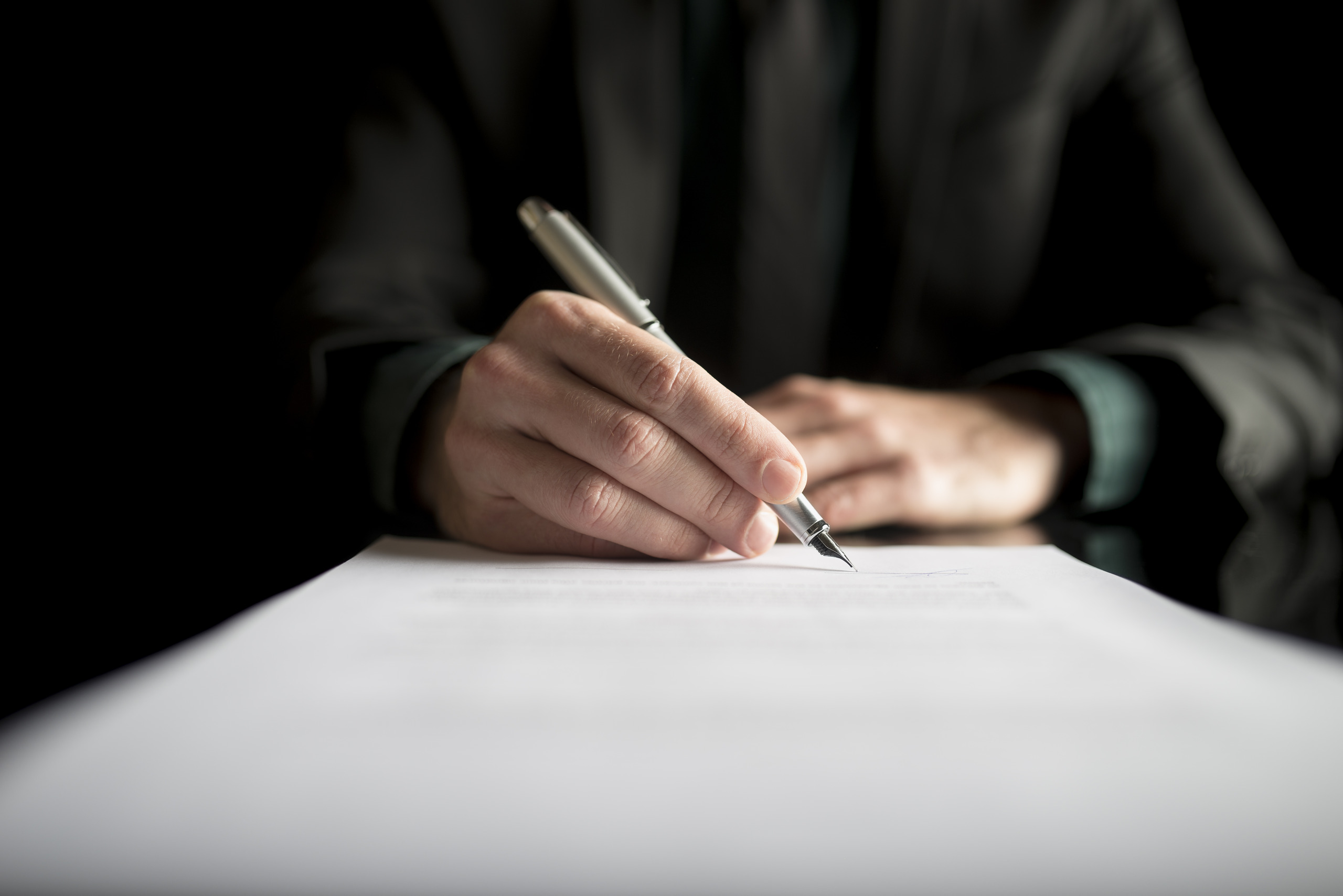 A low-angle view of a person in a dark suit holding a silver fountain pen over a white legal document against a black background, representing suspicious estate plan modifications evaluated under the scope of what is undue influence in a Florida will.